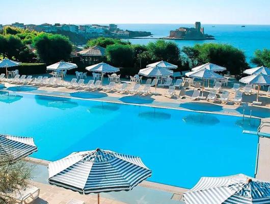 Piscina azzurra con vista mare e castello, ombrelloni a righe, atmosfera rilassante.