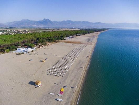 Spiaggia vasta e serena, ombrelloni ordinati, mare calmo e montagne in lontananza.