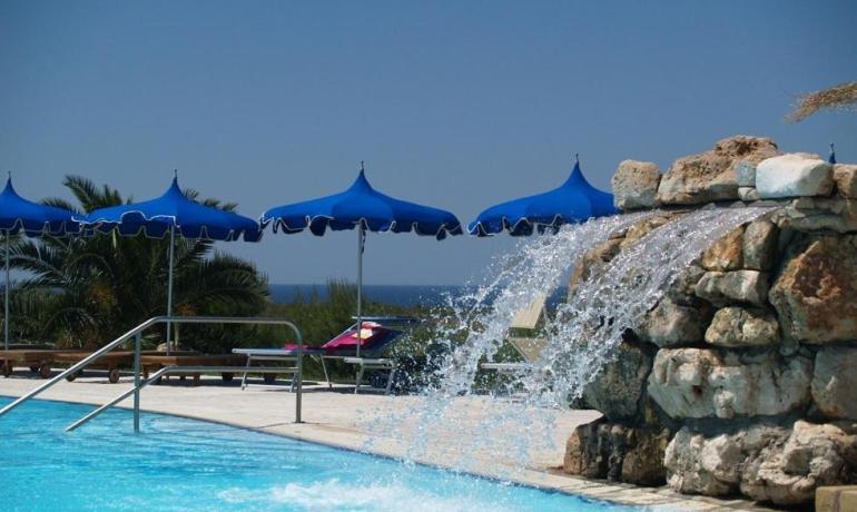 Piscina con cascate, ombrelloni blu e cielo sereno, atmosfera rilassante e vacanziera.