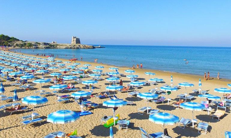 Spiaggia affollata, ombrelloni blu, mare cristallino, torre in lontananza, estate vivace.