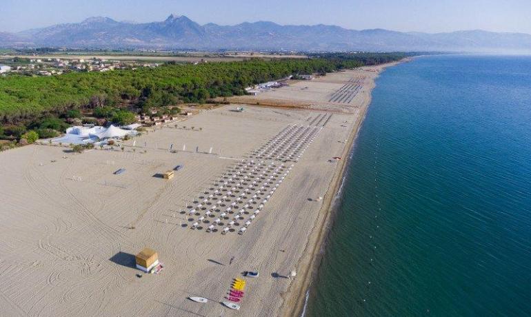 Spiaggia vasta e serena, ombrelloni ordinati, mare calmo e montagne in lontananza.