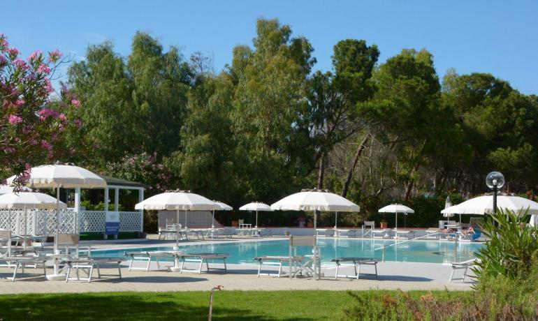 Piscina tranquilla con ombrelloni bianchi, circondata da alberi e fiori colorati.